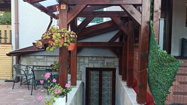 Un espace extérieur abrité, doté d’un mur en pierre, d’une pergola en bois, de jardinières suspendues orn ées de feuillage verdoyant et d’une porte donnant sur un espace intérieur.