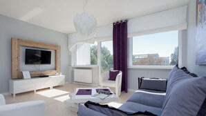 A modern living room with a TV, white furniture, a sofa, and large windows.