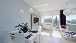 A modern dining area with a white table set for two, a potted plant, and a flat-screen TV mounted on the wall.