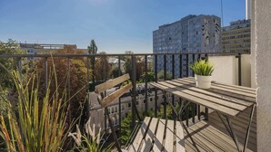 A balcony with a wooden table and a potted plant, overlooking a cityscape with buildings and trees.