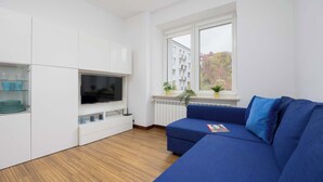 A modern living room with a blue sofa, a flat-screen TV, white cabinetry, and a window with a view of buildings and trees.