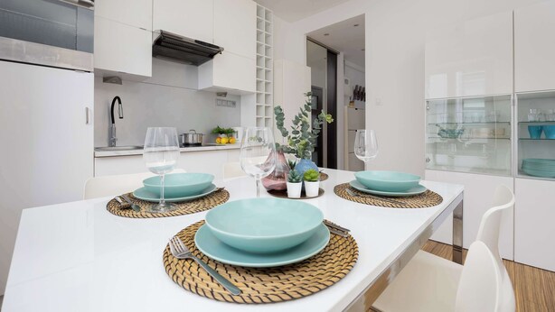 A modern kitchen with white cabinetry, a built-in oven, and a dining area with a white table set for two with teal plates and glassware.