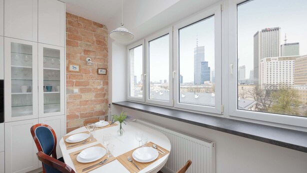 Un comedor moderno con pared de ladrillo, armarios blancos y ventanas grandes con vistas a un paisaje urbano.