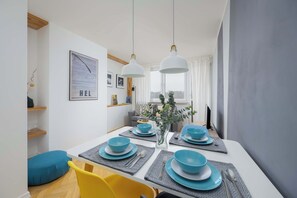 Uma área de refeições moderna com uma mesa branca posta para quatro pessoas, louças azuis e um vaso com folhagens.