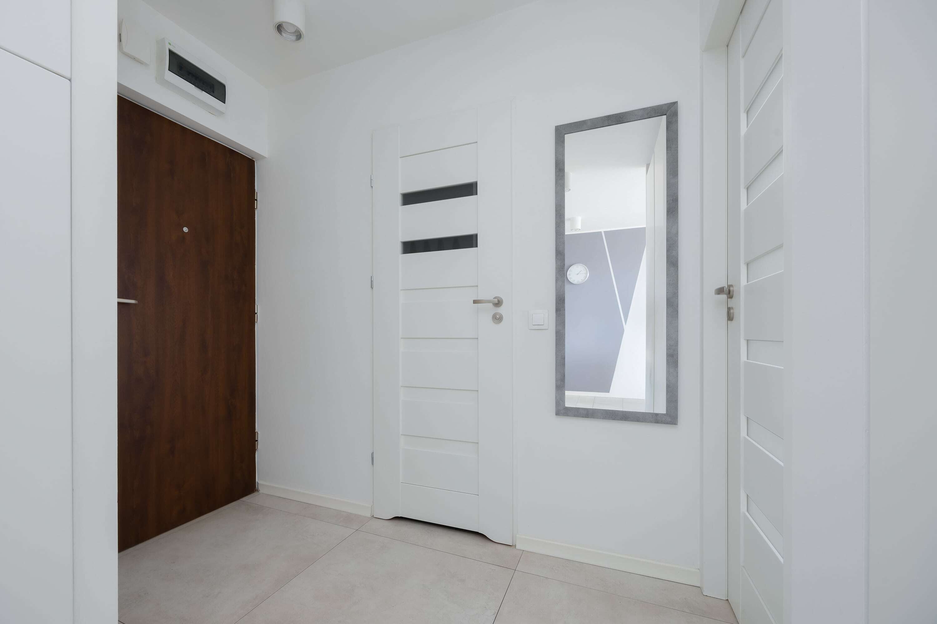 Un pasillo con una puerta de madera, una puerta blanca con listones horizontales y un espejo en la pared.