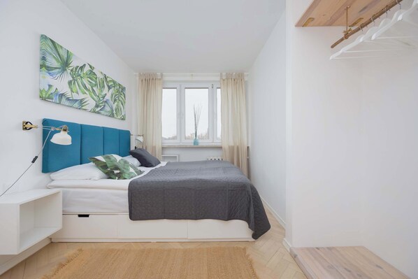 Un dormitorio con cama, mesita de noche, lámpara de pared, ventana con cortinas y un cuadro.