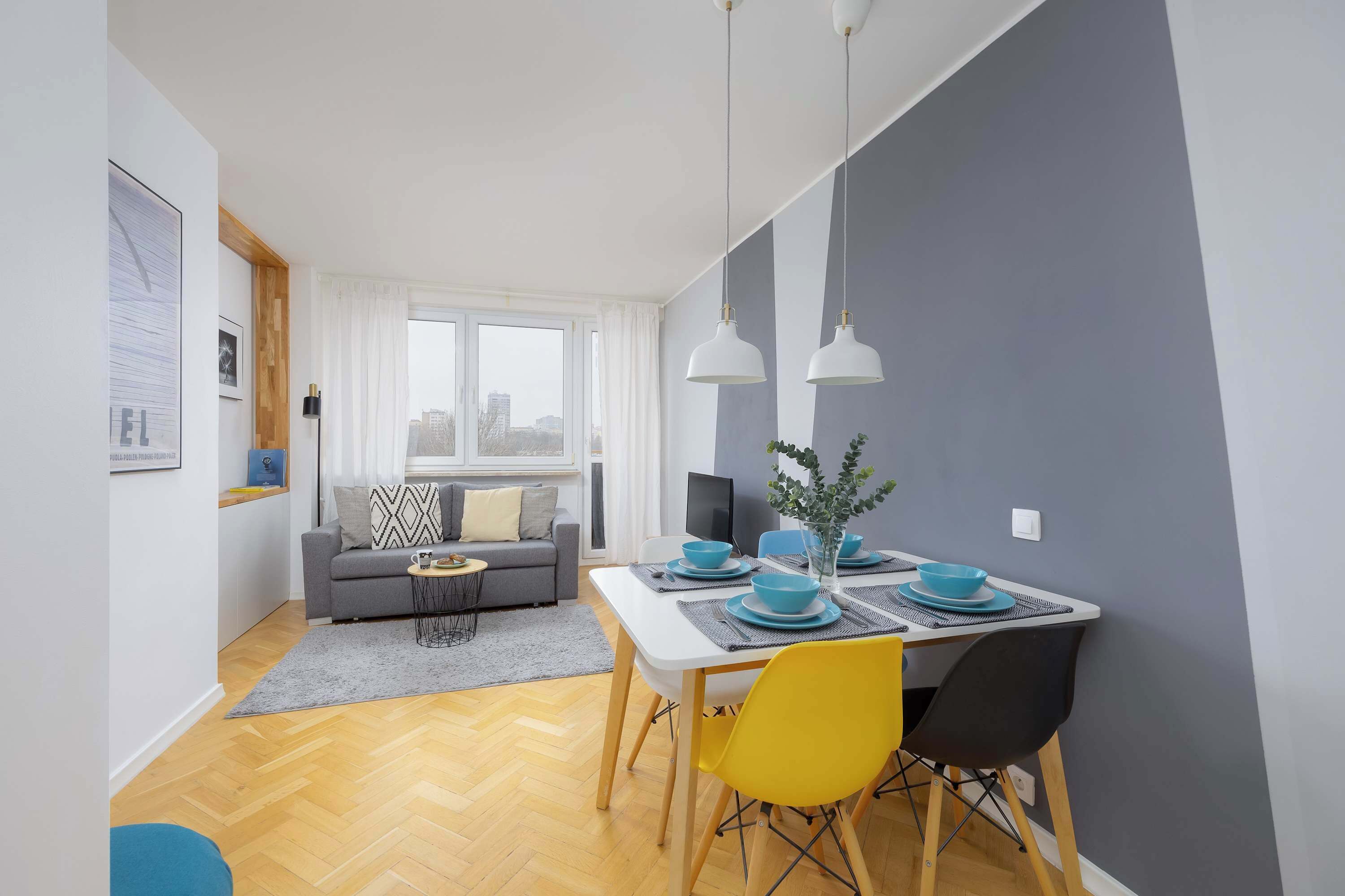 A modern living room with a dining area, featuring a grey sofa, a dining table with blue plates, and a city view through the window.