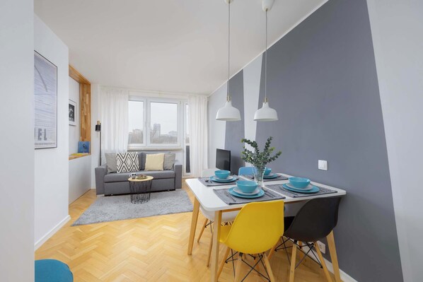 A modern living room with a dining area, featuring a grey sofa, a dining table with blue plates, and a city view through the window.