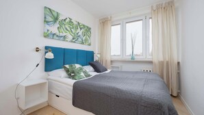 A bedroom with a bed, a nightstand, a wall-mounted lamp, a window with curtains, and a tropical leaf painting.