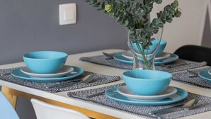 Una mesa de comedor dispuesta con cuencos y platos azules, rodeada de sillas con patas de madera y un florero con follaje verde.