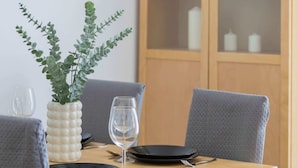 A dining table set with plates, cutlery, and a glass of water. There is a plant in a white vase on the table, and a wooden cabinet in the background.