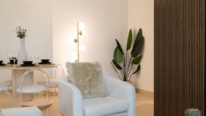 A modern living room with a white sofa, a dining table with chairs, a plant, and a fruit basket.