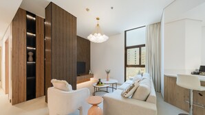 A modern living room with a sofa, armchair, and a round coffee table. There is a TV mounted on the wall and a large window with curtains.