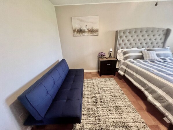 A bedroom with a blue sofa, a bed with striped bedding, a nightstand with a lamp and flowers, and a painting on the wall.
