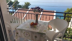A balcony with a table set for two, a bowl of fruit, and a view of the sea.