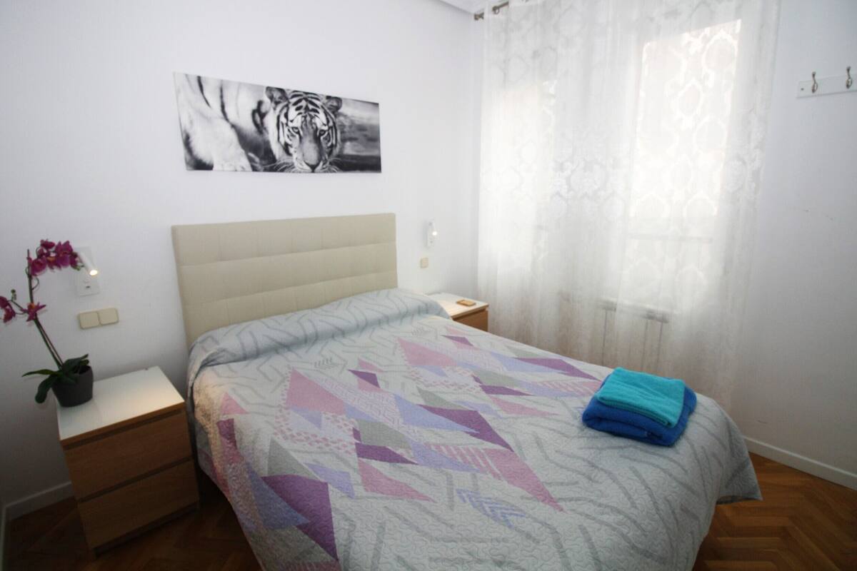 Une chambre à coucher avec un lit, une table de chevet, une plante en pot et une œuvre d’art représentant un tigre accrochée au mur.