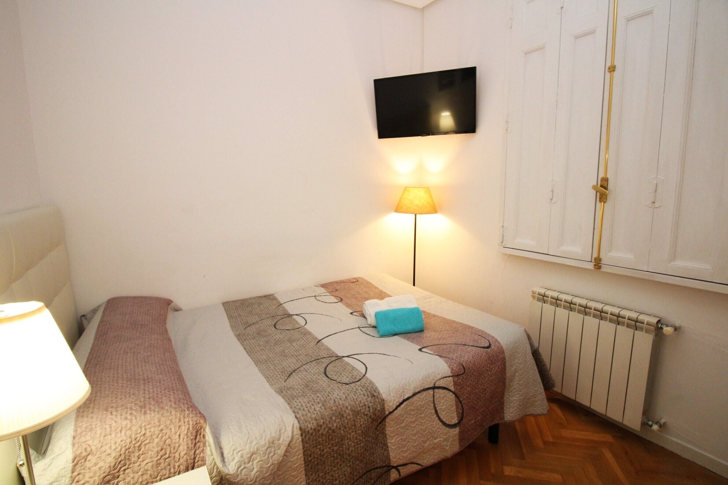 Un dormitorio con cama, lámpara de noche, televisor montado en la pared y radiador.