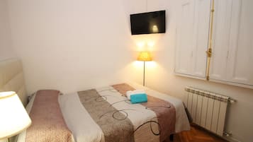 Un dormitorio con cama, lámpara de noche, televisor montado en la pared y radiador.
