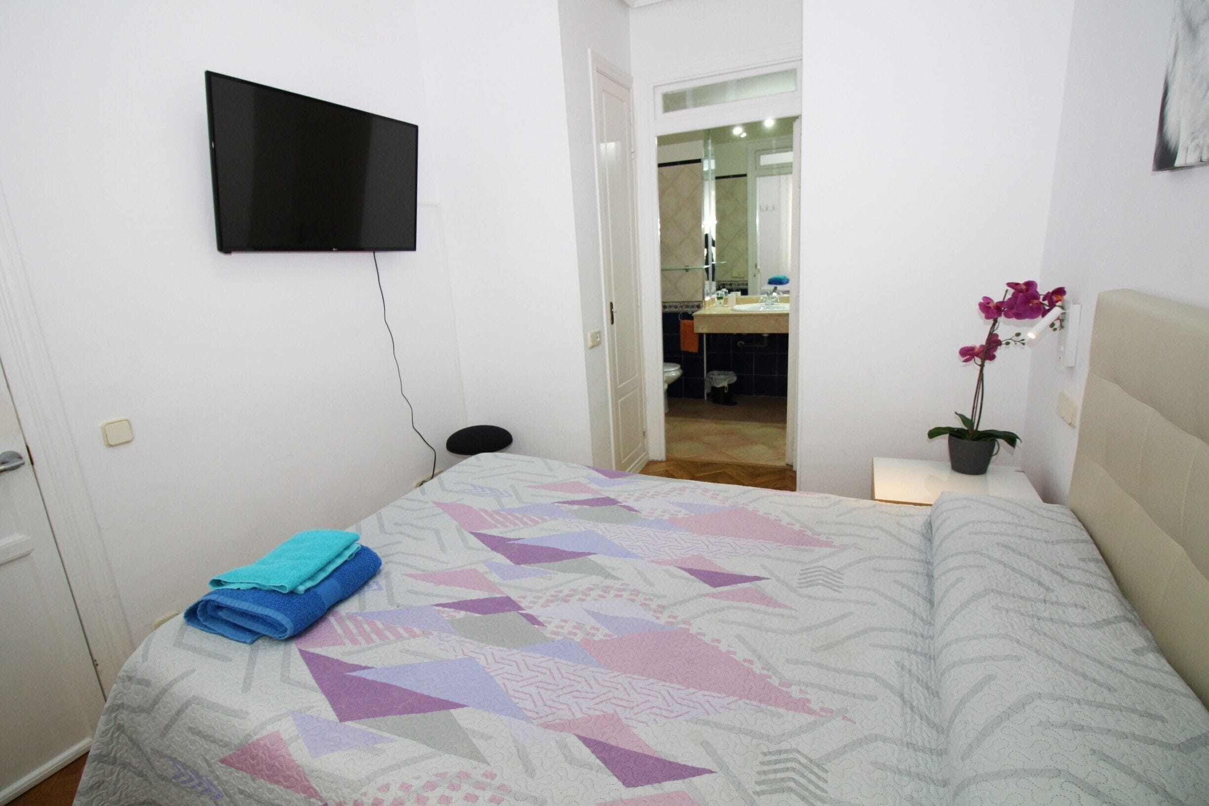 A bedroom with a bed, a TV mounted on the wall, a small bedside table with a plant, and a bathroom visible through an open door.