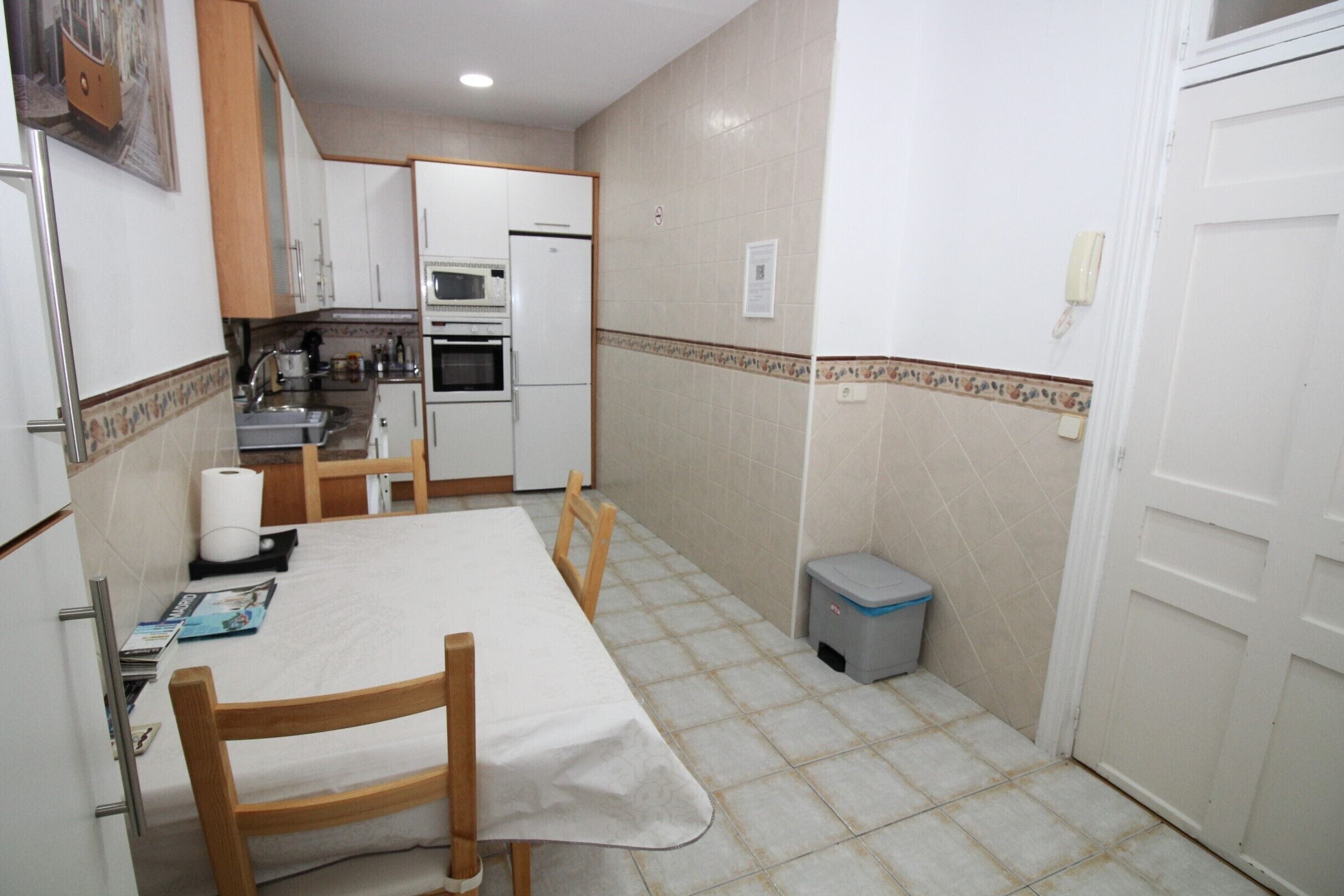A compact kitchen with white cabinets, a microwave, and a refrigerator. There is a dining table with chairs and a trash bin.