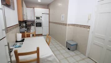 Cocina pequeña con armarios blancos, microondas y refrigerador. Hay una mesa de comedor con sillas y un bote de basura.