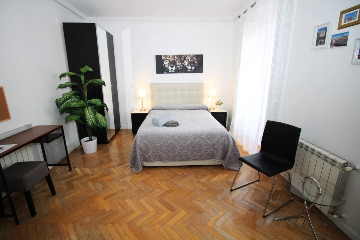 Une chambre à coucher comprenant un lit, des tables de chevet, un bureau, une chaise, une plante en pot et un radiateur.