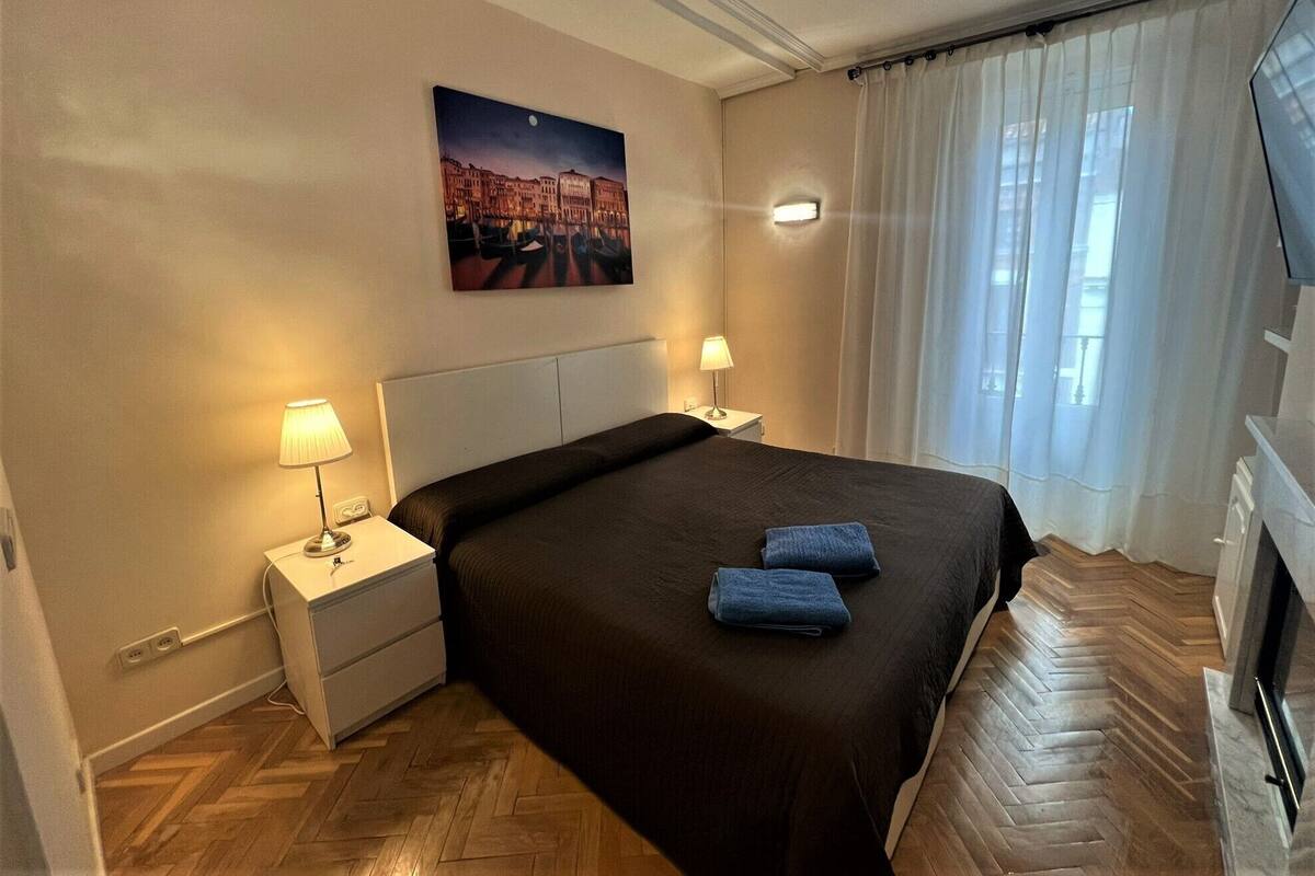 Une chambre d’hôtel avec un grand lit, deux lampes de chevet, une table de nuit, un tableau au mur et une vue sur la ville à travers de fines rideaux.