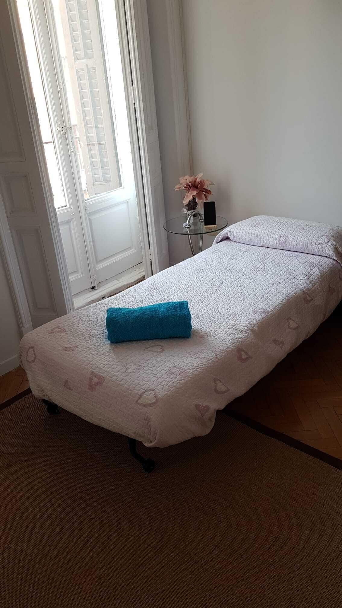 Una cama individual con colcha blanca y toalla azul, una mesita con un florero y un teléfono, y una ventana con persianas.