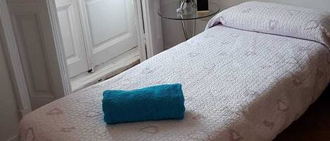 Una cama individual con colcha blanca y toalla azul, una mesita con un florero y un teléfono, y una ventana con persianas.