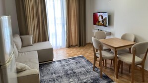 A modern living room with a sofa, a dining table with chairs, a flat-screen TV mounted on the wall, and a large window with curtains. - Palm Suites Apartaments (Vlore)