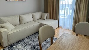 A living room with a beige sofa, two chairs, a wooden coffee table, a patterned rug, a painting on the wall, and a window with curtains. - Palm Suites Apartaments (Vlore)