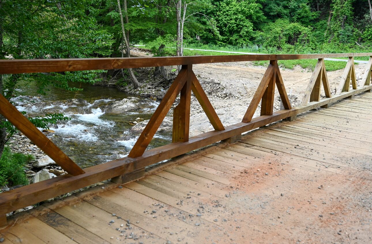 A wooden bridge with a metal railing over a river, surrounded by greenery.