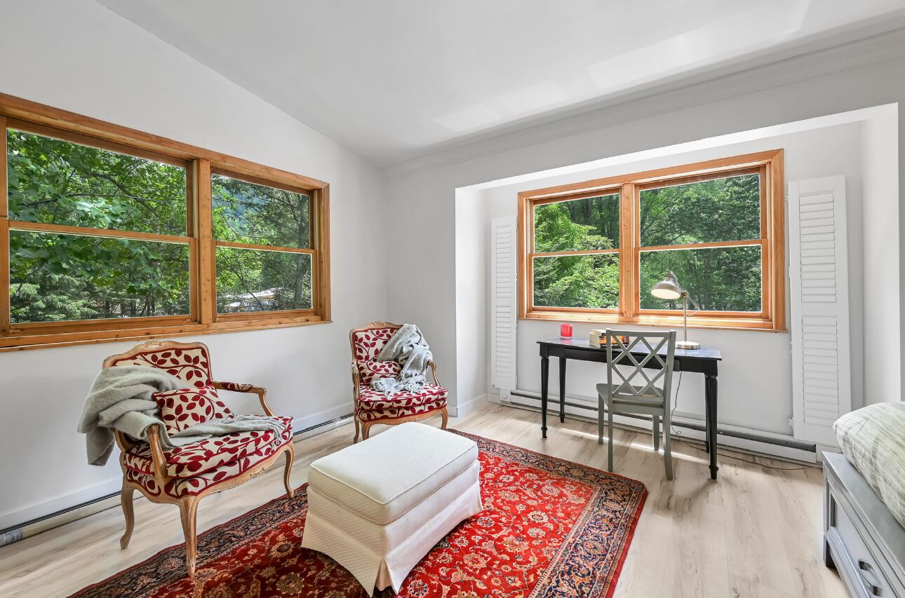 Una habitación con dos ventanas grandes, un escritorio, dos sillas, un tapete rojo con estampado y un reposapiés blanco.