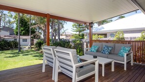 Une terrasse extérieure couverte, aménagée avec un canapé et des fauteuils, une terrasse en bois et offrant une vue sur un quartier résidentiel avec des maisons et des arbres.