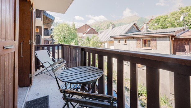 A balcony with a table and chairs, a wooden door, and a view of surrounding buildings and hills.