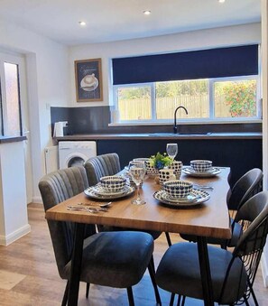 A dining area with a table set for six, chairs, a window with a view, a kitchen area with a washing machine, and a framed picture on the wall.