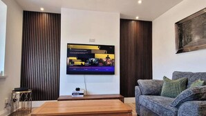 Een moderne woonkamer met een flatscreen televisie aan de muur, een grijze bank met groene kussens en een houten salontafel.