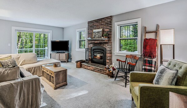 A living room with a fireplace, a television, and two windows with a view of trees.
