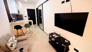 Una sala de estar moderna con un televisor de pantalla plana montado en la pared, un sofá con un cojín estampado, una pequeña mesa de centro negra y una zona de cocina con microondas y hervidor.