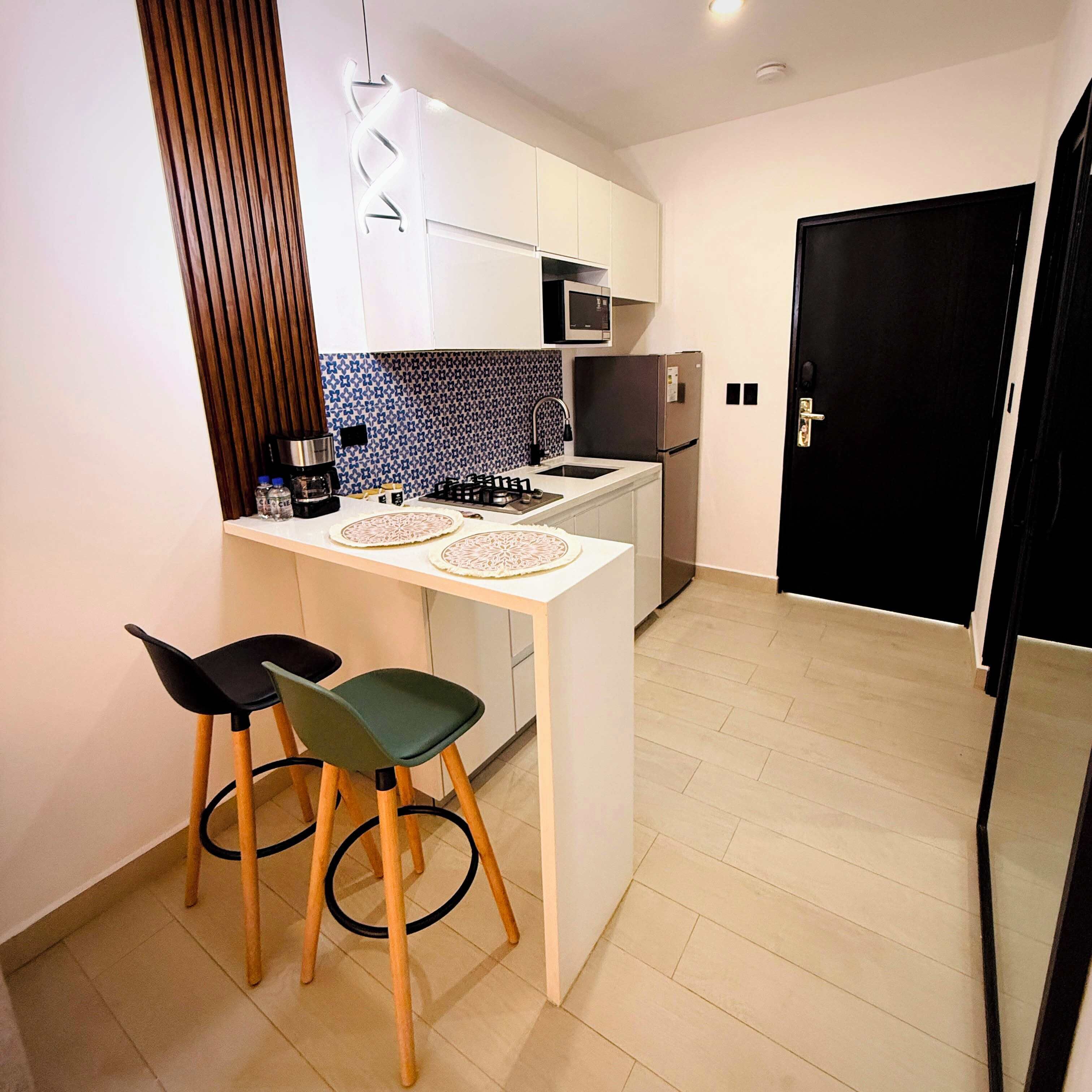 Cocina compacta con una isla central, dos taburetes de bar, un microondas, un refrigerador y una puerta negra.