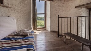 Une pièce rustique avec un banc en bois, un lit recouvert d’une couverture à rayures et une grande fenêtre donnant sur un paysage verdoyant.