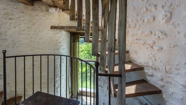 Un escalier rustique avec des marches en bois et une rampe en métal, menant à un espace extérieur où l’on peut apercevoir de la verdure à travers une fenêtre.