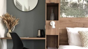 Un dormitorio con cama, escritorio con silla, espejo y vista a árboles por la ventana.