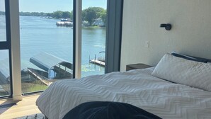 A bedroom with a large bed, a view of a lake with boats, and a glass door leading to an outdoor area. - Lake Minnetonka Retreat (Mound)