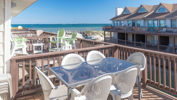 Une terrasse dotée d’une table à plateau en verre et de chaises blanches, offrant une vue sur une plage et un immeuble à multiples fenêtres.