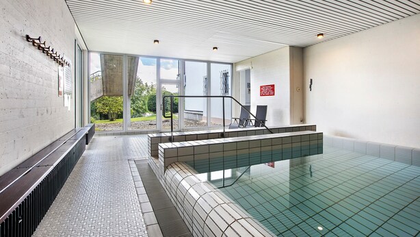 En swimmingpool med flisegulv, en bænk og et glasgelænder.