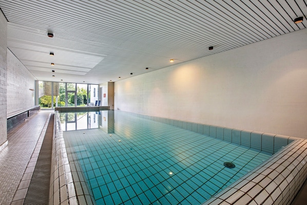 An indoor swimming pool with a tiled floor, a sloped ceiling with recessed lighting, and a view of greenery outside.