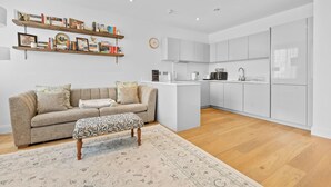 A modern living room with a sofa, ottoman, and a rug. There is a kitchen area with white cabinets and a countertop. - London Road Apartment in Isleworth (Isleworth)