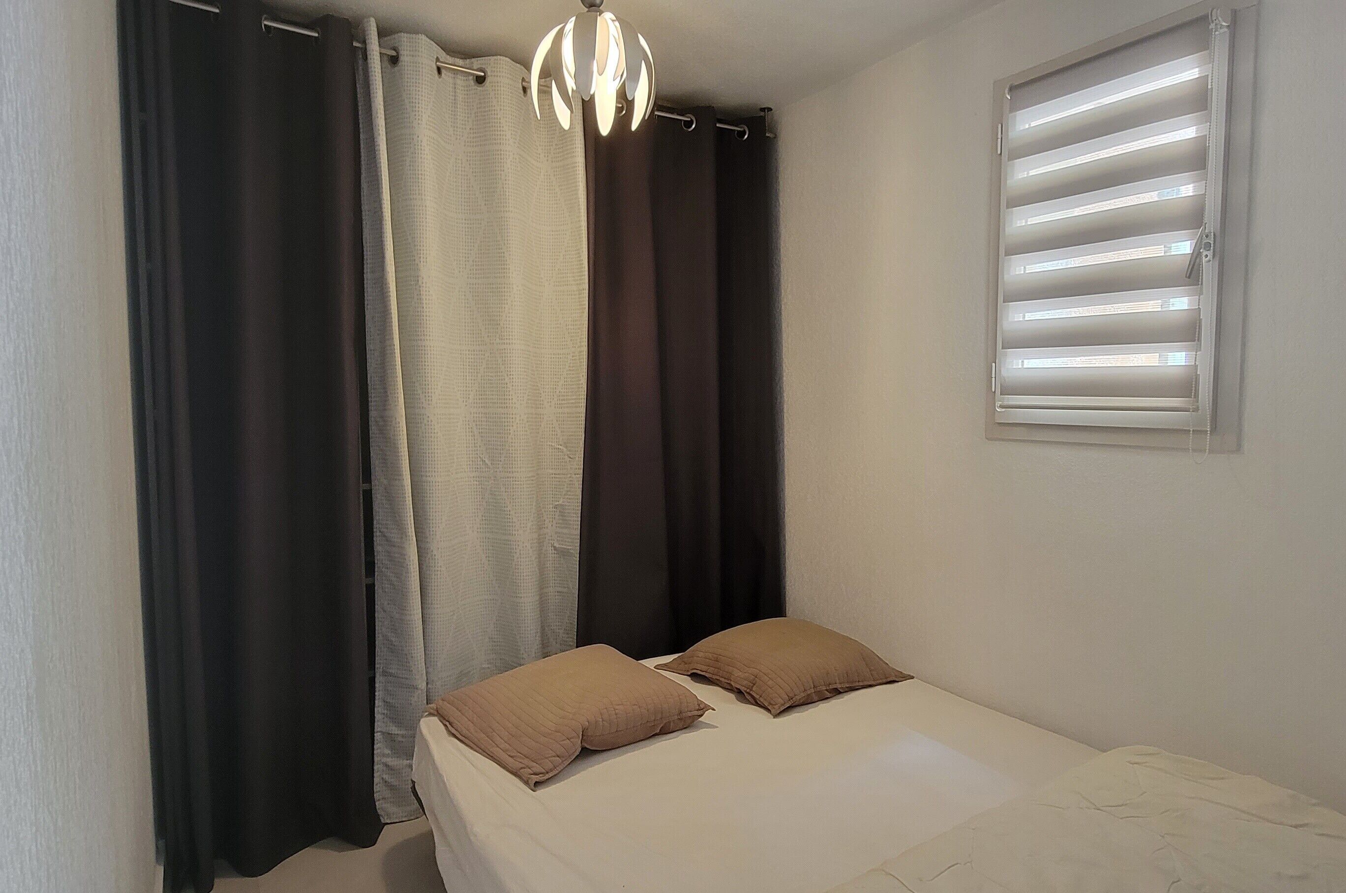 Et soveværelse med en seng, to puder, en loftlampe og et vindue med persienner.