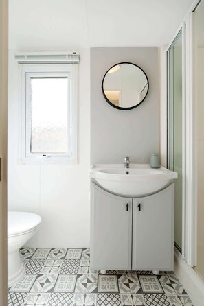 A bathroom with a white sink, a round mirror, a toilet, and patterned floor tiles. - The Chester-2 bed Static Caravan (Jedburgh)